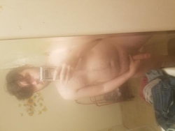 A person with a curvy build takes a mirror selfie in a bathroom, topless and holding a phone, with visible body hair