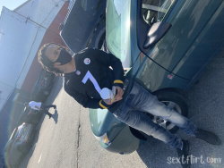 A person in a black Pittsburgh Steelers jersey and jeans leans against a green car, wearing a face mask and holding a