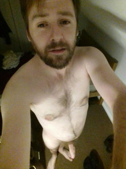 A shirtless man with a beard takes a selfie in a dimly lit room, showing his bare torso and lower body.