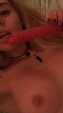Blonde woman with bare chest holding a red toy in her mouth, wearing a black choker necklace.