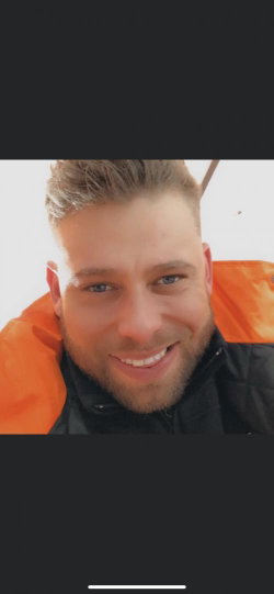 Man with short styled hair and beard, wearing an orange life vest over a black jacket, smiling at the camera.