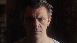 A man with graying hair and a serious expression, wearing a light-colored shirt, in a dimly lit indoor setting.