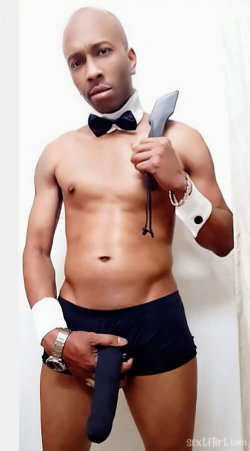 Bald Black man with muscular build wearing bow tie and white cuffs, holding a black dildo in one hand and a gray object