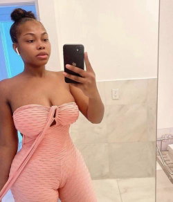 A curvy woman with dark skin takes a mirror selfie in a pink textured bodysuit, hair in a bun, standing in a tiled
