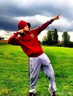 A young man in a red hoodie and gray sweatpants poses outdoors on a grassy field under stormy skies, pointing upward