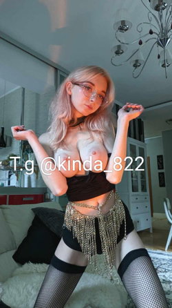 Blonde woman with glasses in a black crop top and fishnet stockings poses indoors, wearing a chain belt and kneeling on