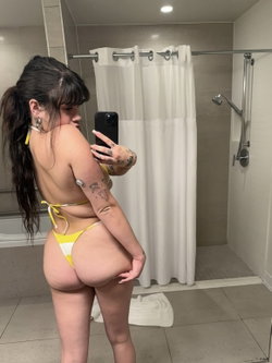 Curvy woman with dark hair in a yellow and white bikini takes a mirror selfie in a bathroom, showing tattoos on her