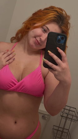 Redheaded woman in a bright pink bikini top takes a mirror selfie in a bathroom, smiling softly with colorful nails.