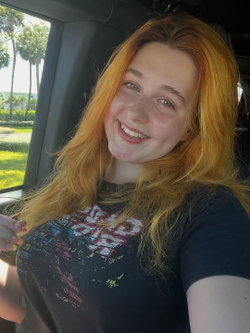 A smiling woman with long red hair and freckles takes a selfie in a car, wearing a dark graphic t-shirt.