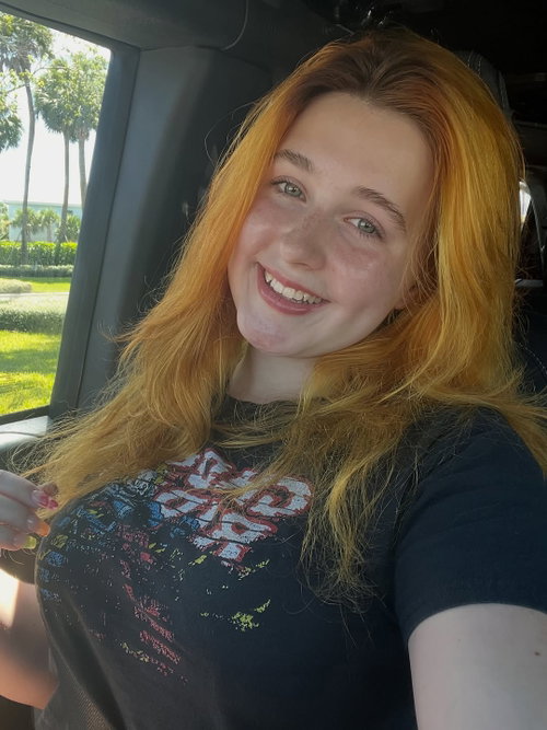 A smiling woman with long red hair and freckles takes a selfie in a car, wearing a dark graphic t-shirt.