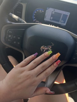 A hand with colorful, patterned nails rests on a Ford Mustang steering wheel inside a car, with legs visible in the