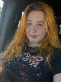 Redheaded woman with freckles and green eyes smiling in a car, wearing a graphic black t-shirt, holding her hair.