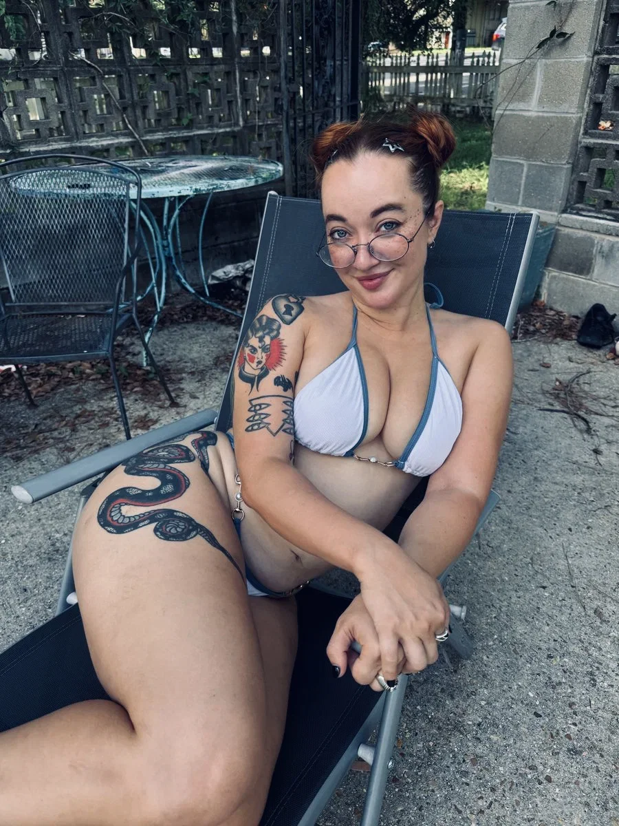 A woman with tattoos and glasses lounges in a bikini on a patio chair, smiling at the camera.