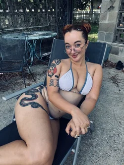 A woman with tattoos and glasses lounges in a bikini on a patio chair, smiling at the camera.