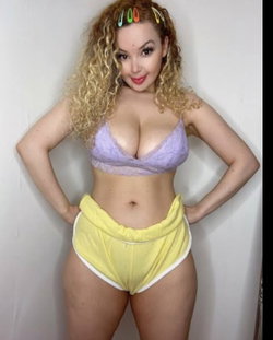 Curvy woman with curly blonde hair wearing a purple lace bra and yellow high-waisted shorts, posing with hands on hips