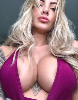 Blonde woman with wavy hair and a purple top, showing a tattooed chest and shoulder in a close-up selfie.