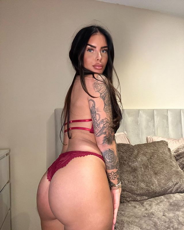 Brunette woman with full figure in red lace lingerie poses in bedroom, showing tattooed arm and back.