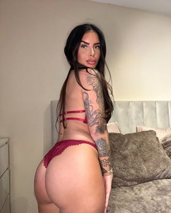 Brunette woman with full figure in red lace lingerie poses in bedroom, showing tattooed arm and back.