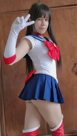 Woman in Sailor Moon cosplay with long dark hair, white gloves, blue pleated skirt, and red boots posing indoors.