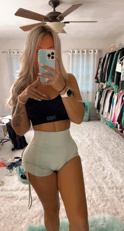 Blonde woman in a black crop top and high-waisted shorts taking a selfie in a bedroom.