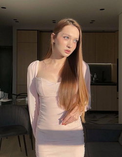 A woman with long light brown hair wears a fitted light pink dress with sheer sleeves, standing in a modern