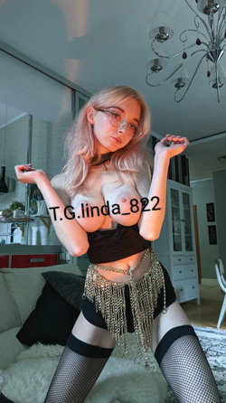Blonde woman with glasses poses in a black crop top, chain belt, and fishnet stockings in a modern living room.