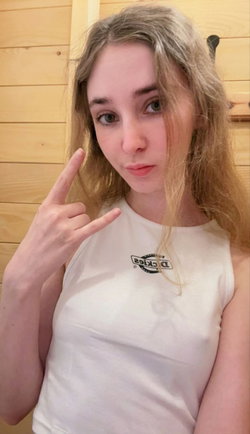 Blonde woman in a white tank top making a hand gesture, standing indoors against a wooden wall.