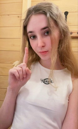 Blonde woman in a white tank top poses with finger raised, standing indoors against a wooden wall.
