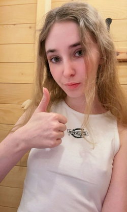 Blonde woman in a white tank top gives a thumbs-up in front of a wooden wall.