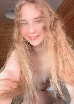 Blonde woman with long wavy hair takes a close-up selfie, bare chest visible beneath her hair, smiling softly indoors.