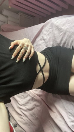 A person lies on a pink bed, wearing a black strappy top with hand resting on hip, long nails and rings visible.