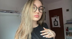 Blonde woman with long hair and round glasses wears a black top, posing indoors with hand on chest in a room with