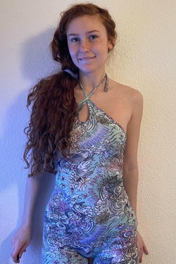 A young woman with curly red hair wears a colorful patterned halter jumpsuit, standing against a plain wall with a