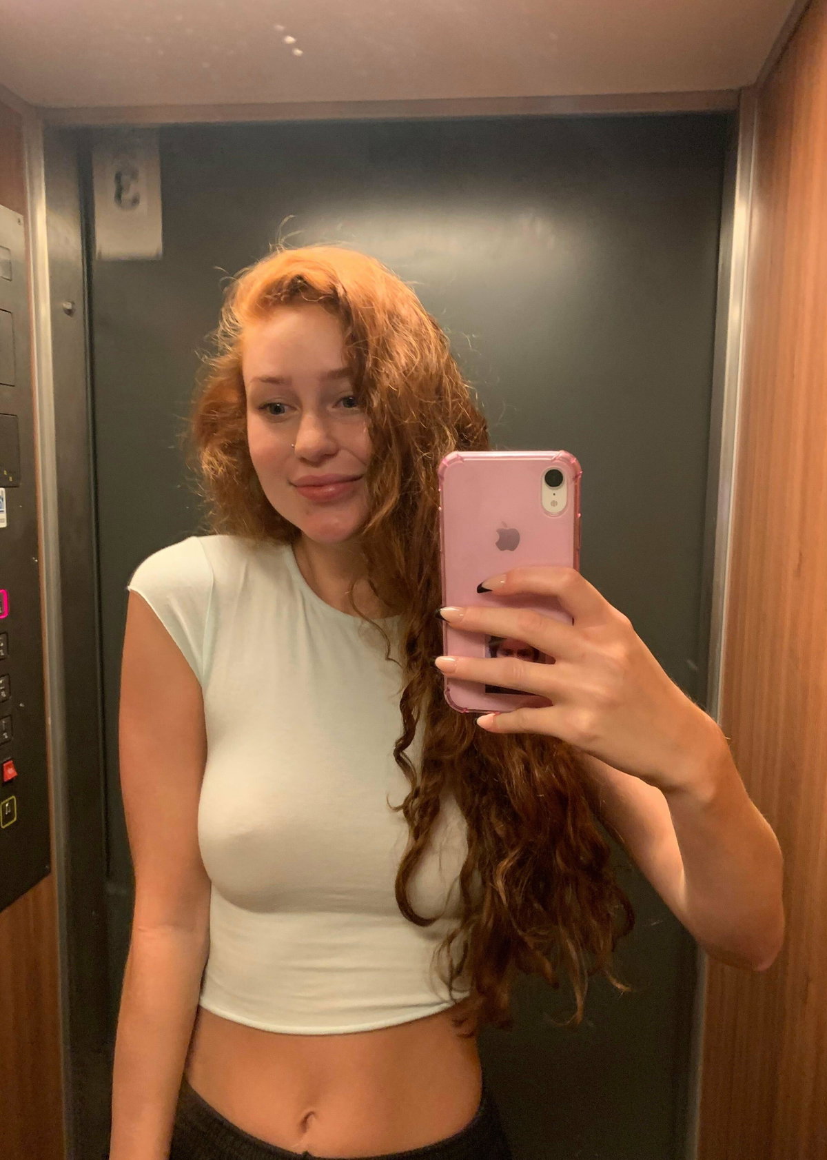 Curvy redhead with long curly hair taking a selfie in an elevator wearing a white crop top.