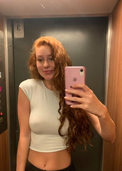 Curvy redhead with long curly hair taking a selfie in an elevator wearing a white crop top.