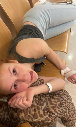 Redheaded woman with freckles lies on a wooden chair, wearing a gray crop top and light blue jeans, resting her head on
