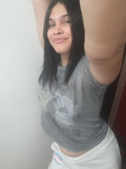 A woman with dark hair smiles while stretching her arms above her head, wearing a gray crop top and white pants against