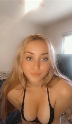 Blonde woman with blue eyes in black lace bra, leaning forward in a bedroom setting.