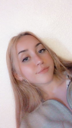 Blonde woman with long hair and light makeup, wearing a gray top, posing against a plain wall in a close-up selfie.