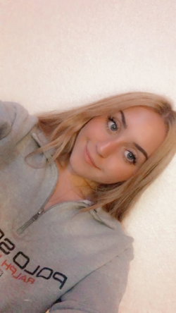 Blonde woman with blue eyes in a gray zip-up hoodie, lying on a light-colored surface and smiling at the camera.