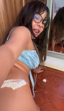 Brunette woman with glasses in a light blue top, posing on a wooden floor with white cream on her lower back, taking a