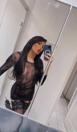 Woman with long black hair wearing sheer black sparkly outfit takes mirror selfie in tiled bathroom.