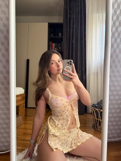 A woman with long brown hair takes a mirror selfie in a floral lace lingerie set, kneeling on a fluffy rug in a bedroom.
