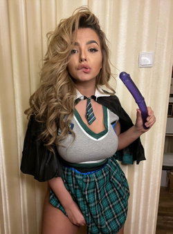 Blonde woman in a schoolgirl outfit with a plaid skirt and tie, holding a purple toy, posing against a cream curtain.