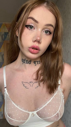 Petite woman with auburn hair, freckles and tattoos wears sheer white lace bra indoors.