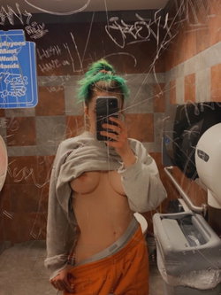 Woman with green hair takes a mirror selfie in a graffiti-filled bathroom while lifting her sweatshirt.