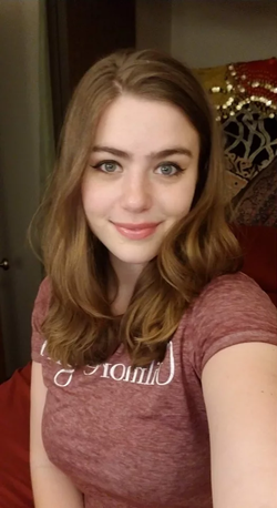 A woman with shoulder-length brown hair and blue eyes smiles softly in a selfie, wearing a maroon t-shirt indoors.