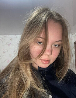 A young woman with long blonde hair and fair skin wears a dark high-neck top, looking directly at the camera in a
