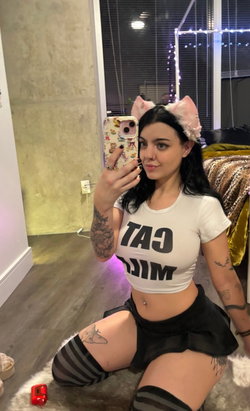 A woman with dark hair and tattoos kneels on a fluffy rug, wearing a white crop top, black skirt, striped thigh-highs,