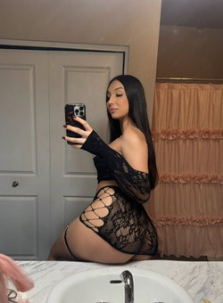 Curvy woman with long dark hair in black lace outfit taking a selfie in a bathroom.